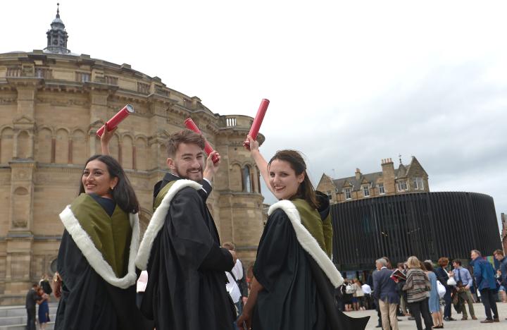Three students celebrate with their families after graduation, holding red scrolls up to the sky.