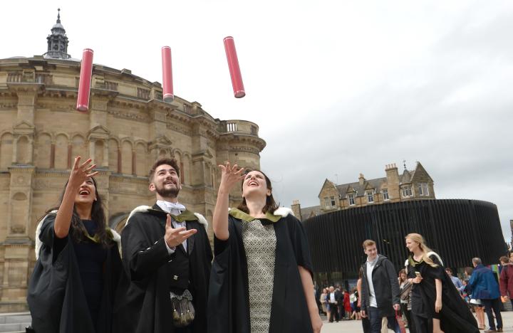 Three graduates standing together and celebrating their graduation by throwing red cylinder tubes in the air