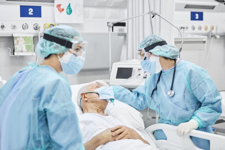 Female doctor speaks to with ICU patient, an older man with a oxygen mask, along with a female coworker in ICU.