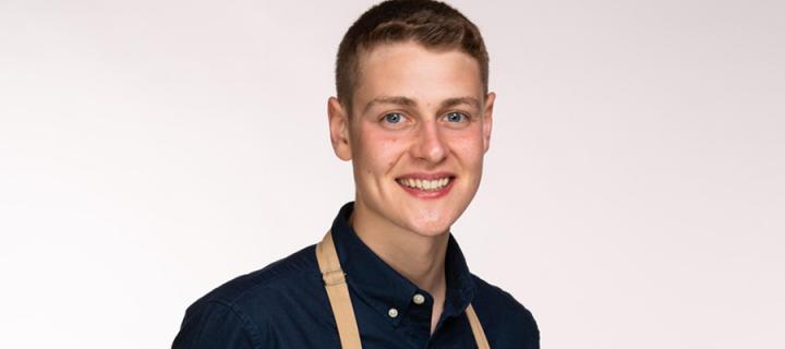Young male, with light brown hair, wearing a blue shirt and a brown apron, looking into the camera and smiling.