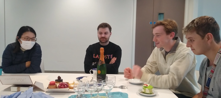 Photo of lab group members seated around a table that has pastries, glasses and a bottle on it