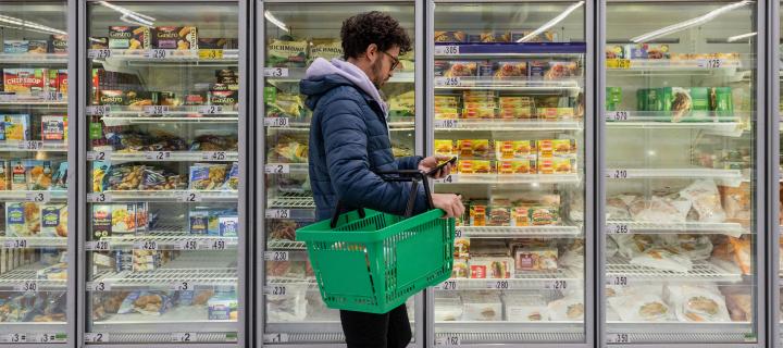 Person shopping in a supermarket, holding a basket and walking down a freezer aisle.