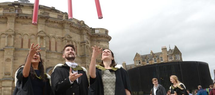 Three graduates standing together and celebrating their graduation by throwing red cylinder tubes in the air
