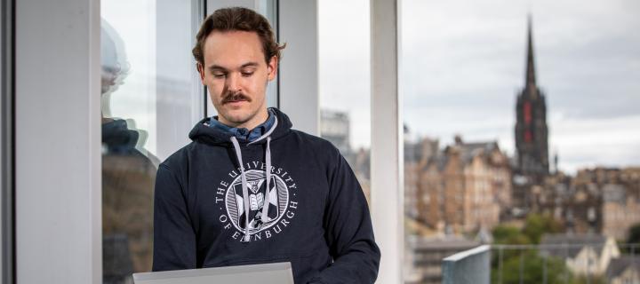 Male student wearing University-branded jumper using laptop with Edinburgh skyline in background