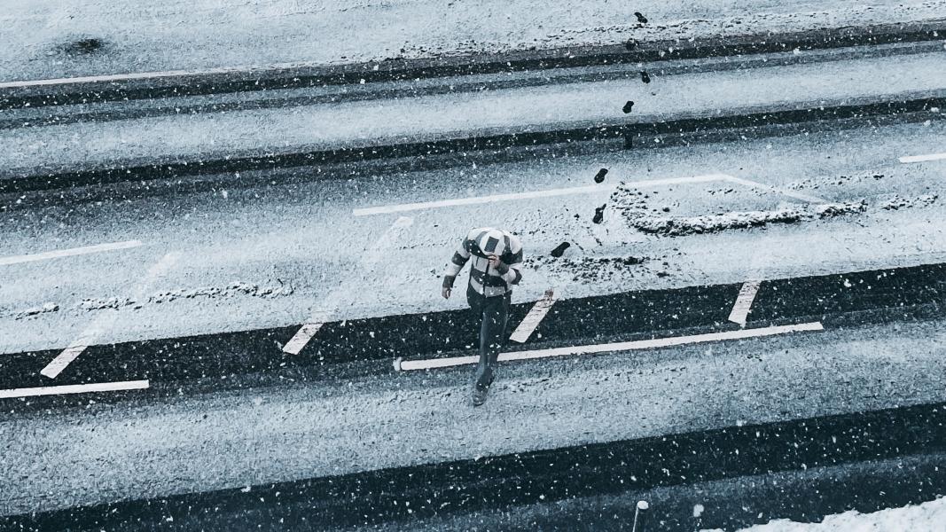 Person holding their hood to shield their face as they walk across a snow covered road while snow falls around them.