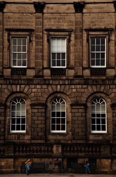 Two people sat on two separate benches in front of a large old building with white window frames.