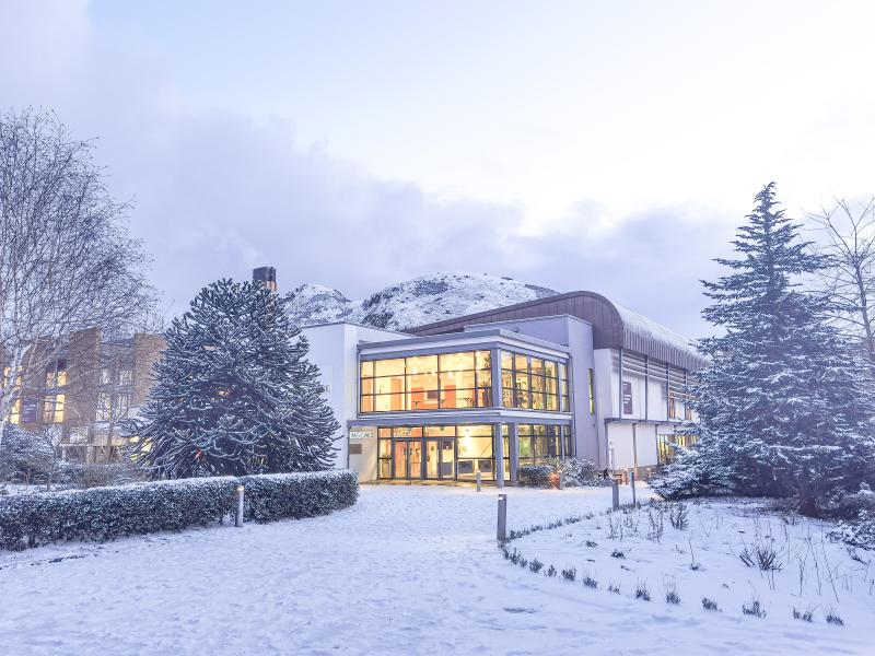 Snow covered building with snow covered hill in the background and coniferous trees in foreground.