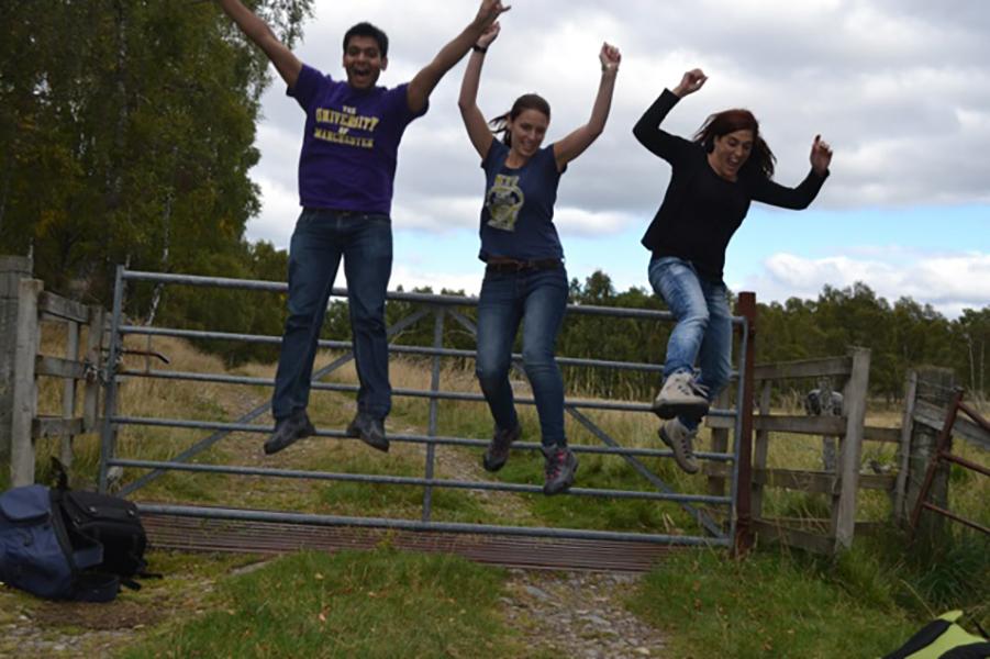 Three group members jump in the air in front of a fence outside in a grassy landscape surrounded by trees