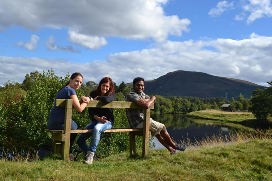 Three lab goup members sit on a bench by a lake, the landscape around is grassy, with trees and a mountain in the background
