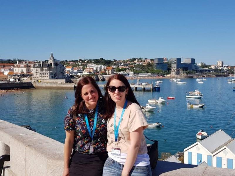 Alba and another lab member sit on a harbour wall which overlooks a costal town and boats in the harbour