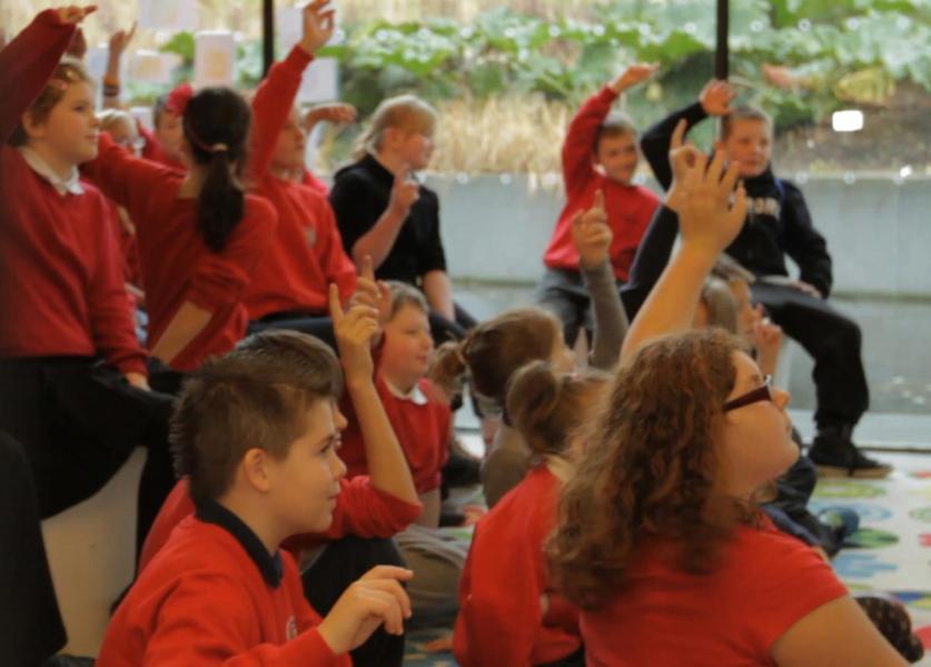 A group of school children sit on the floor with hands raised